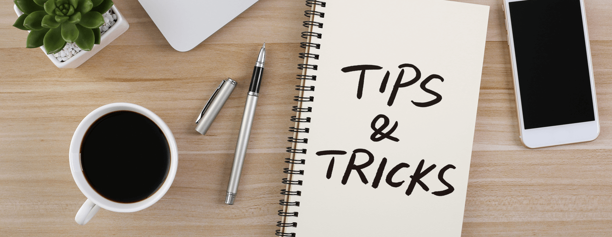 Notebook on wooden desk that says tips and tricks, along with coffee cup, pens, a plant, and a smart phone