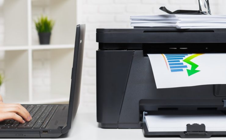 A person typing on laptop computer next to a multifunction printer on table