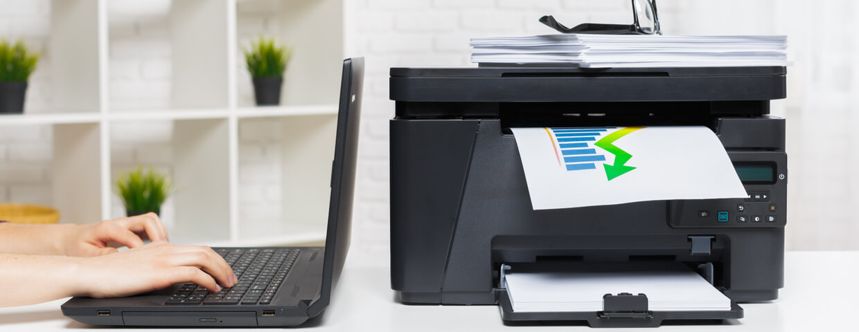 A person typing on laptop computer next to a multifunction printer on table