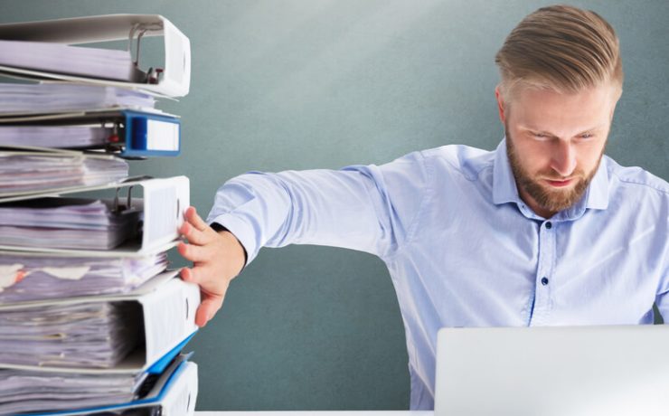 Man seated looking at his laptop and using one hand to hold up a stack of full binders
